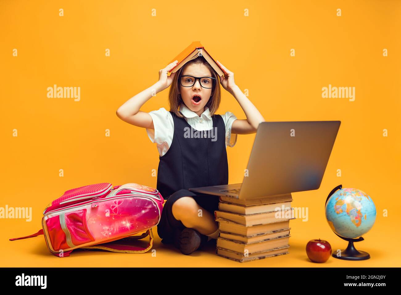 Surprised schoolgirl sits behind a stack of books and laptop holds book ...