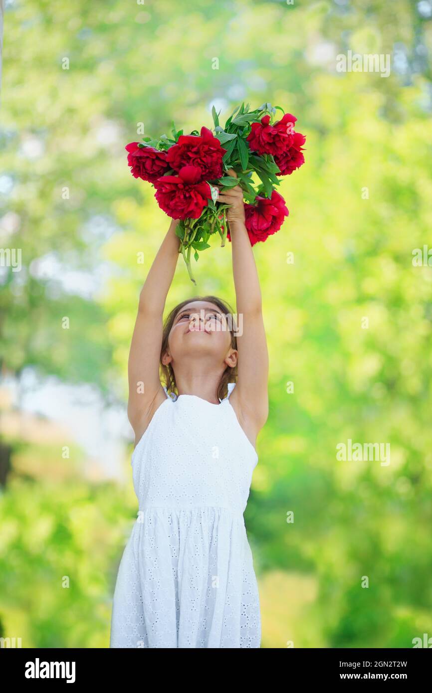 Little girl with peony flowers. Child wearing white dress playing in a summer garden. Kids gardening. Children play outdoors. Toddler kid with flower Stock Photo