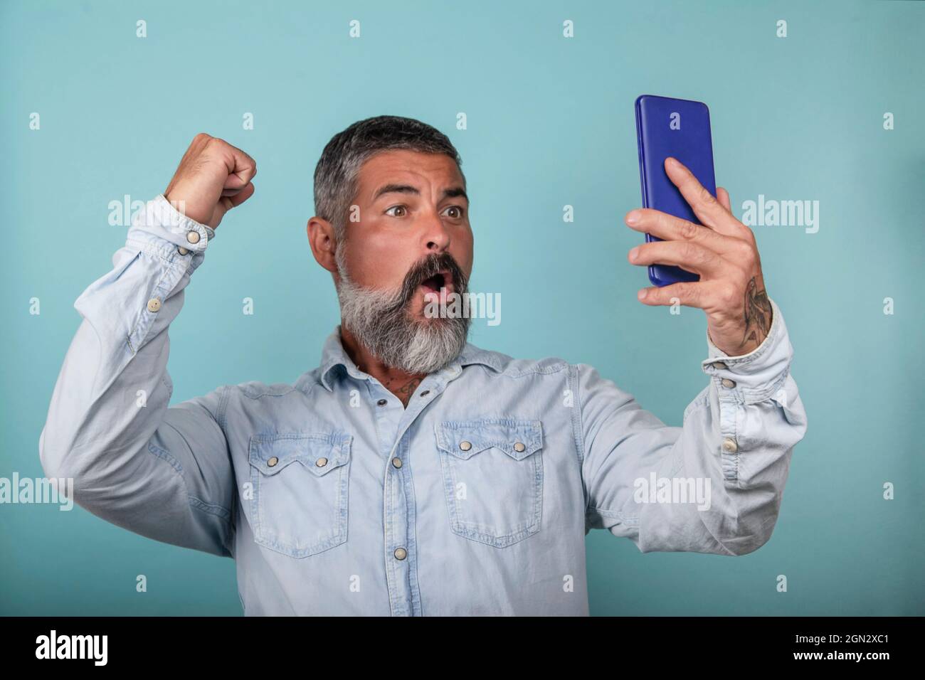 Photo of happy man triumphing, holding mobile phone and making winner gesture standing over light blue background Stock Photo