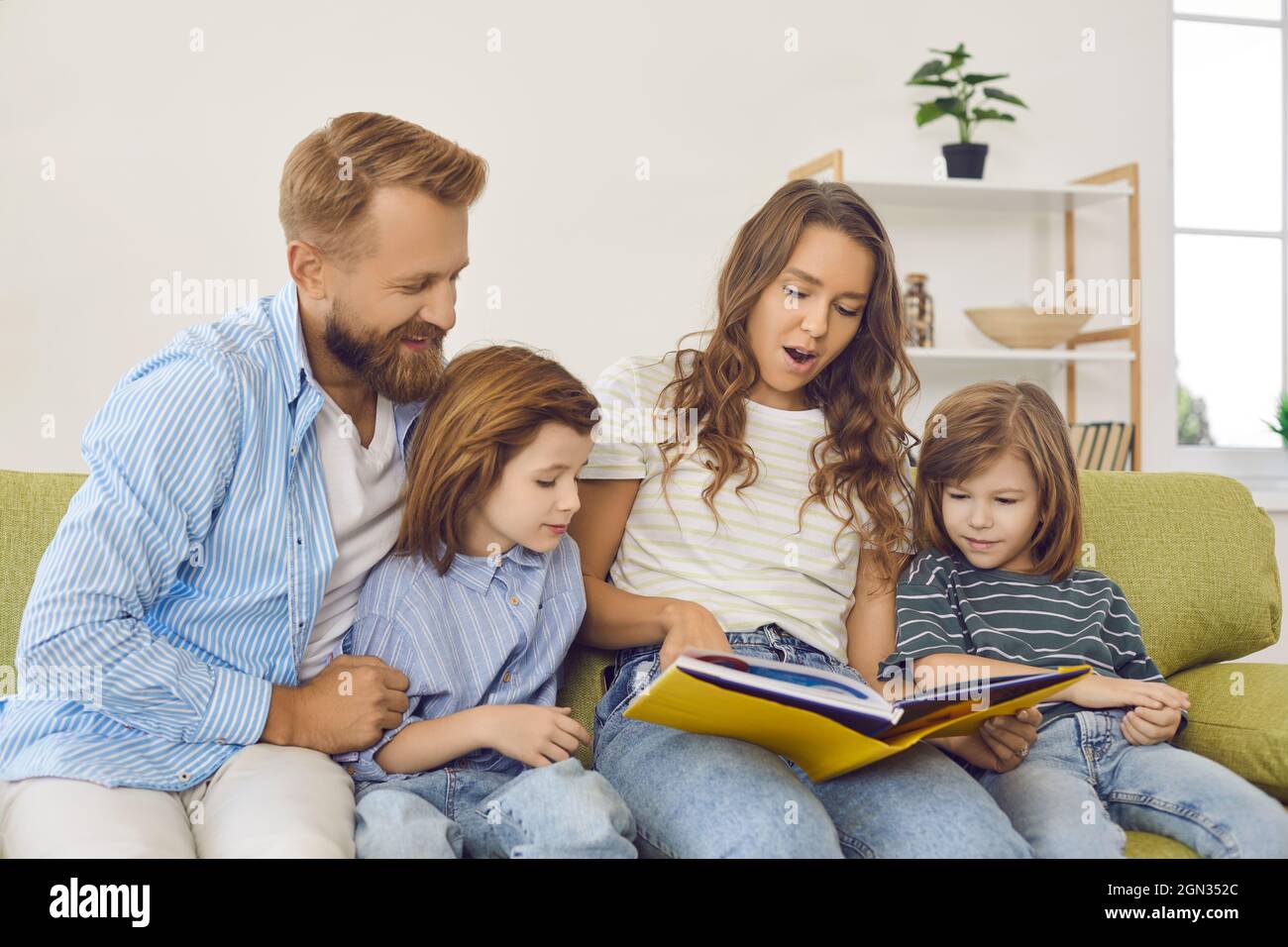 Parents and little children reading book of stories while sitting on couch at home Stock Photo