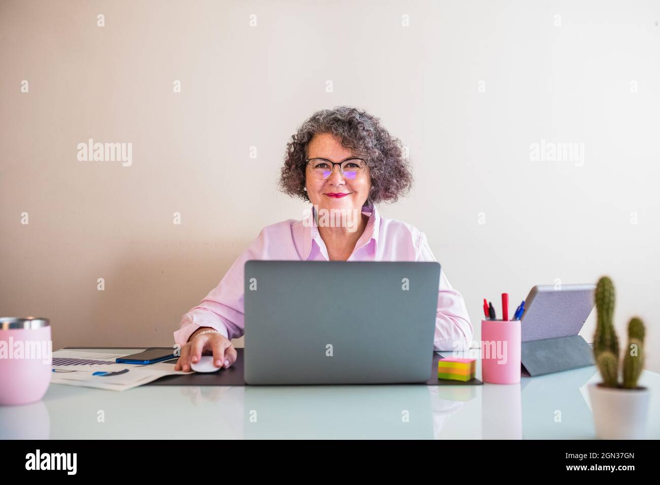 Cheerful senior businessWoman in eyewear sitting at table with netbook and mouse while looking at camera on light background Stock Photo