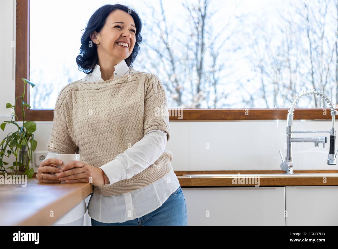 Mature confident Woman standing looking away in kitchen near countertop holding mug of hot