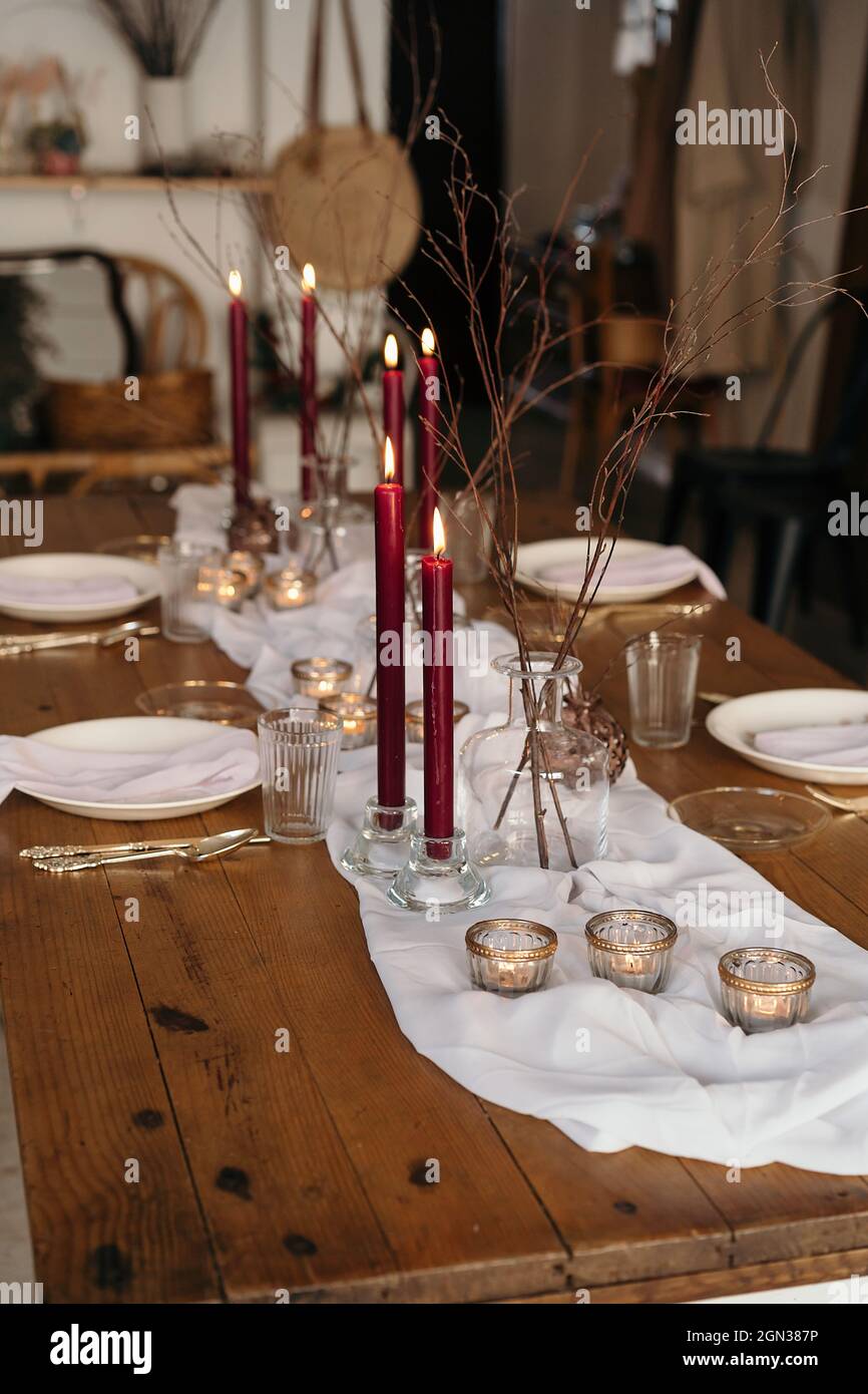 White tablecloth and plates placed on festive table decorated with burning candles and dry branches of tree Stock Photo