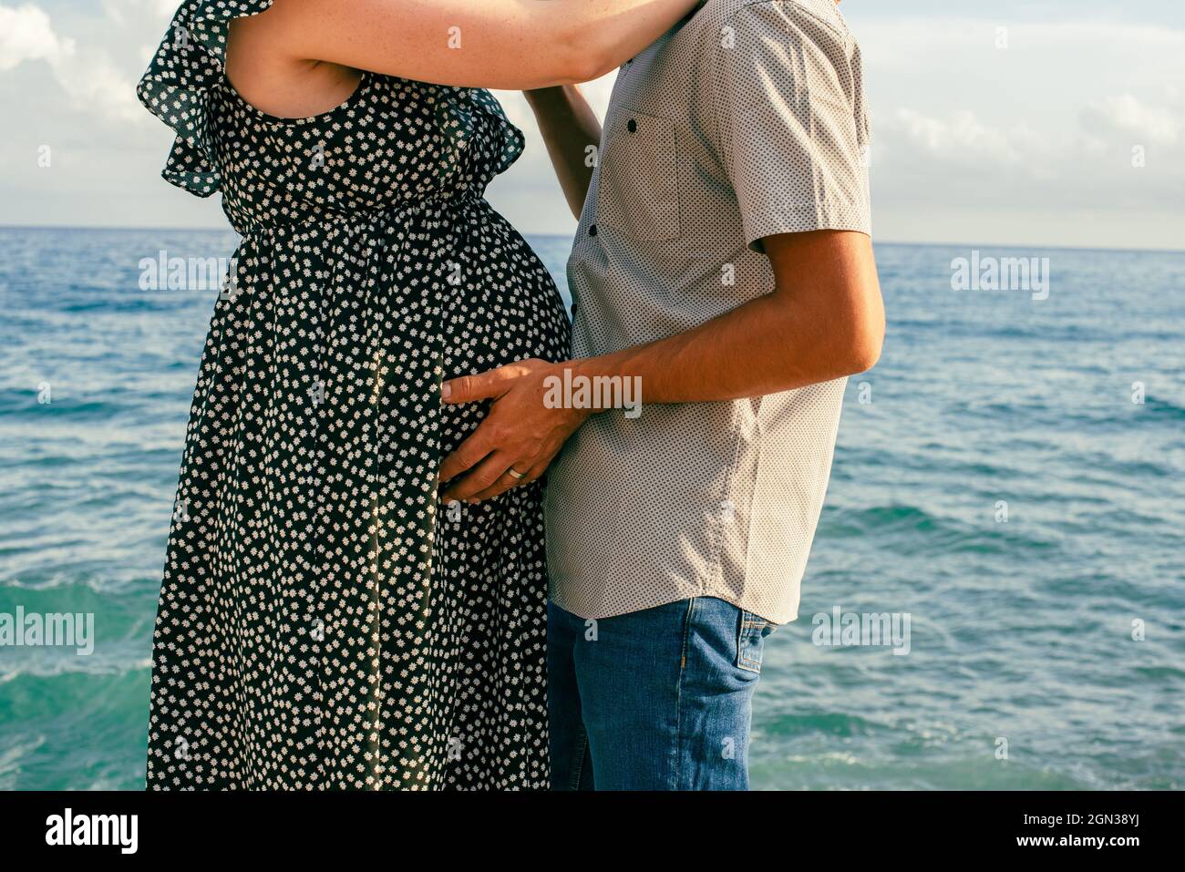 Pregnant woman hugging her partner by the sea. Interracial couple with pregnant expecting a baby. Stock Photo
