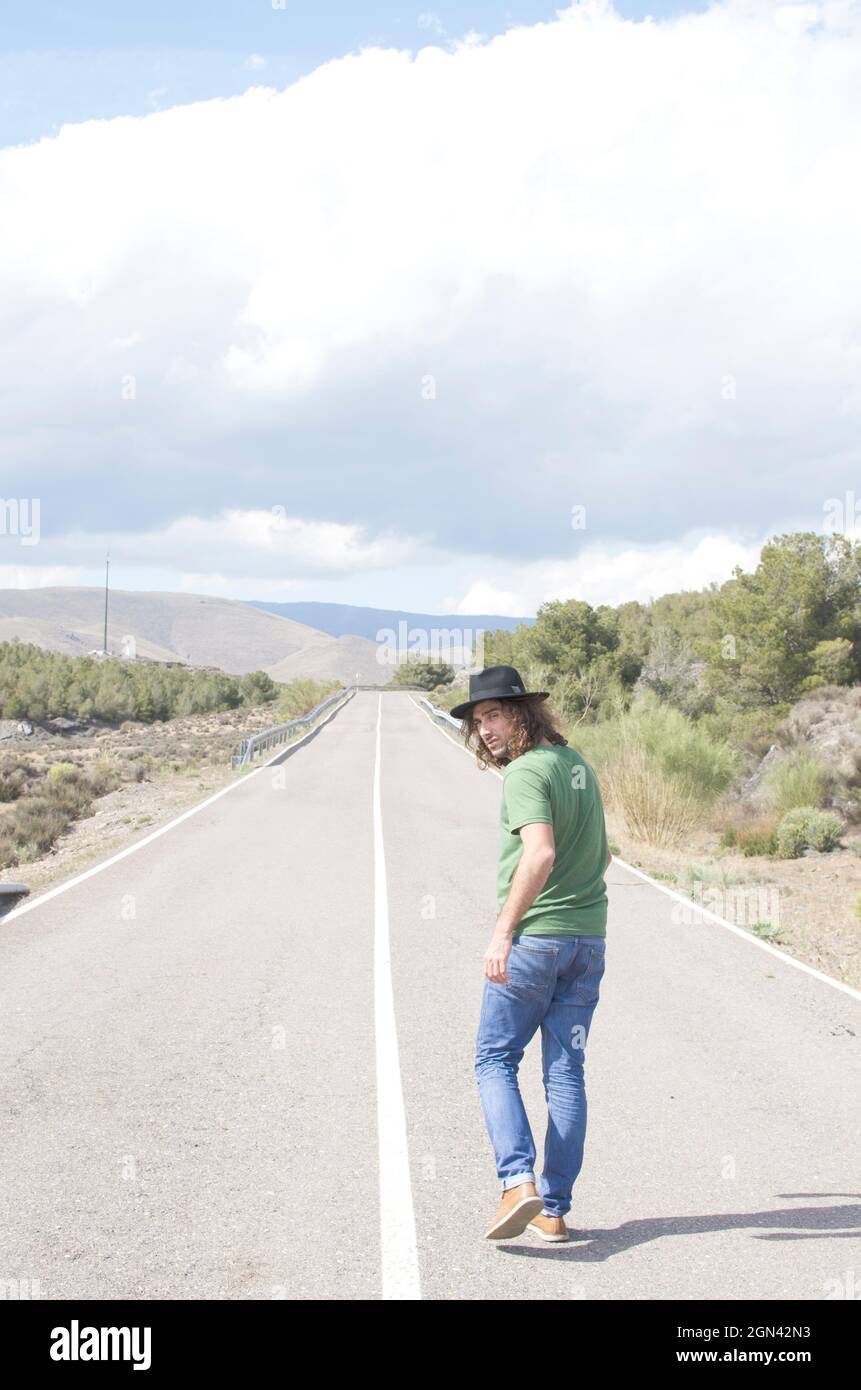 young man in hat walking down a deserted road turns to look Stock Photo