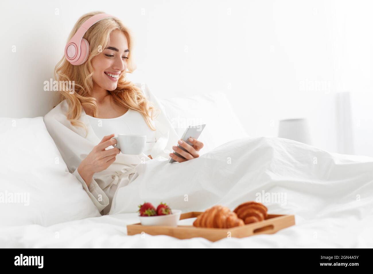 Smiling millennial pretty european blonde woman in robe and headphones reading news, chatting, watch video Stock Photo