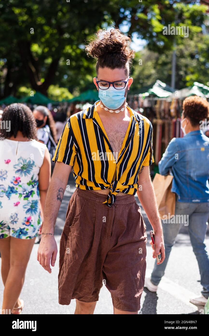 A stylish gay man wearing wearing a surgical face mask, yellow and black striped shirt, brown linen shorts, and choker necklace made with buzio conche Stock Photo