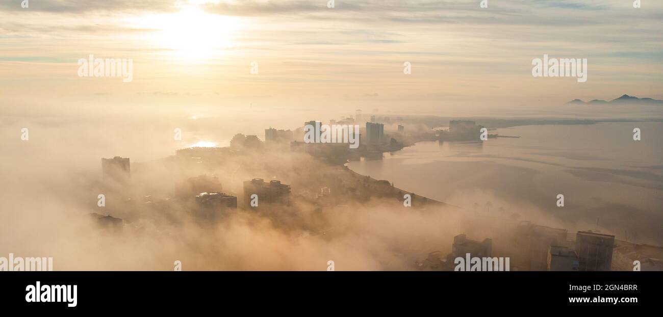 Aerial drone point of view La Manga del Mar Menor cityscape and Mediterranean sea during sunrise, glowing sun on cloudy misty sky, picturesque scenery Stock Photo