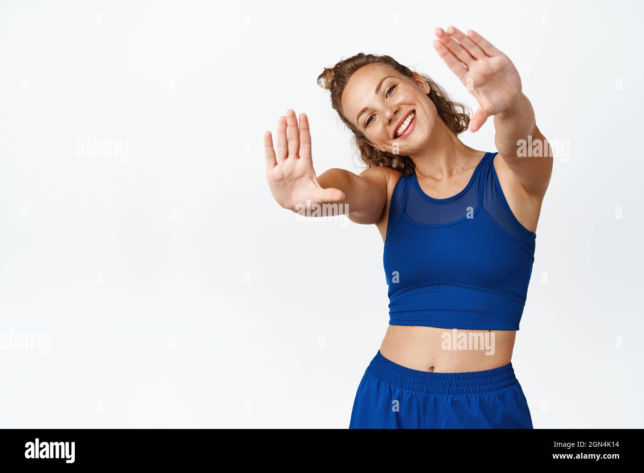 Happy sporty girl in fitness clothing, stretching out hands and smiling, picturing moment, saving memory, standing against white background Stock Photo