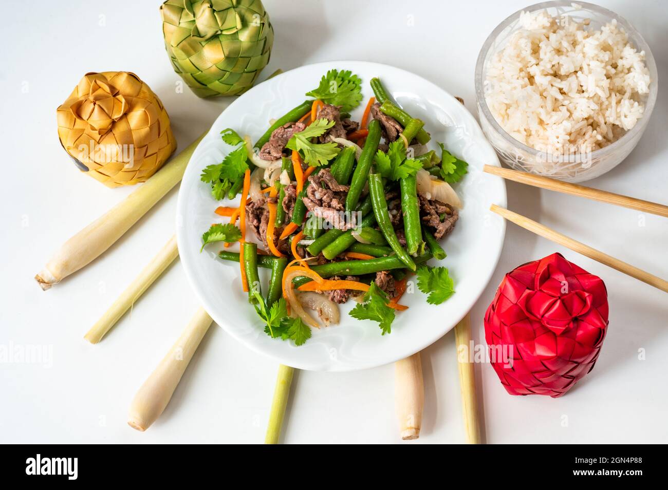 Food background with vietnamese meal, plate with fried beef with green bean and rice, lemon grass, chopstick and 3 color bamboo box on white backgroun Stock Photo