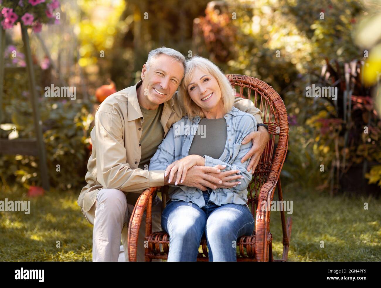 Loving elderly spouses resting in countryside, woman sitting in wicker chair, man embracing wife, copy space Stock Photo