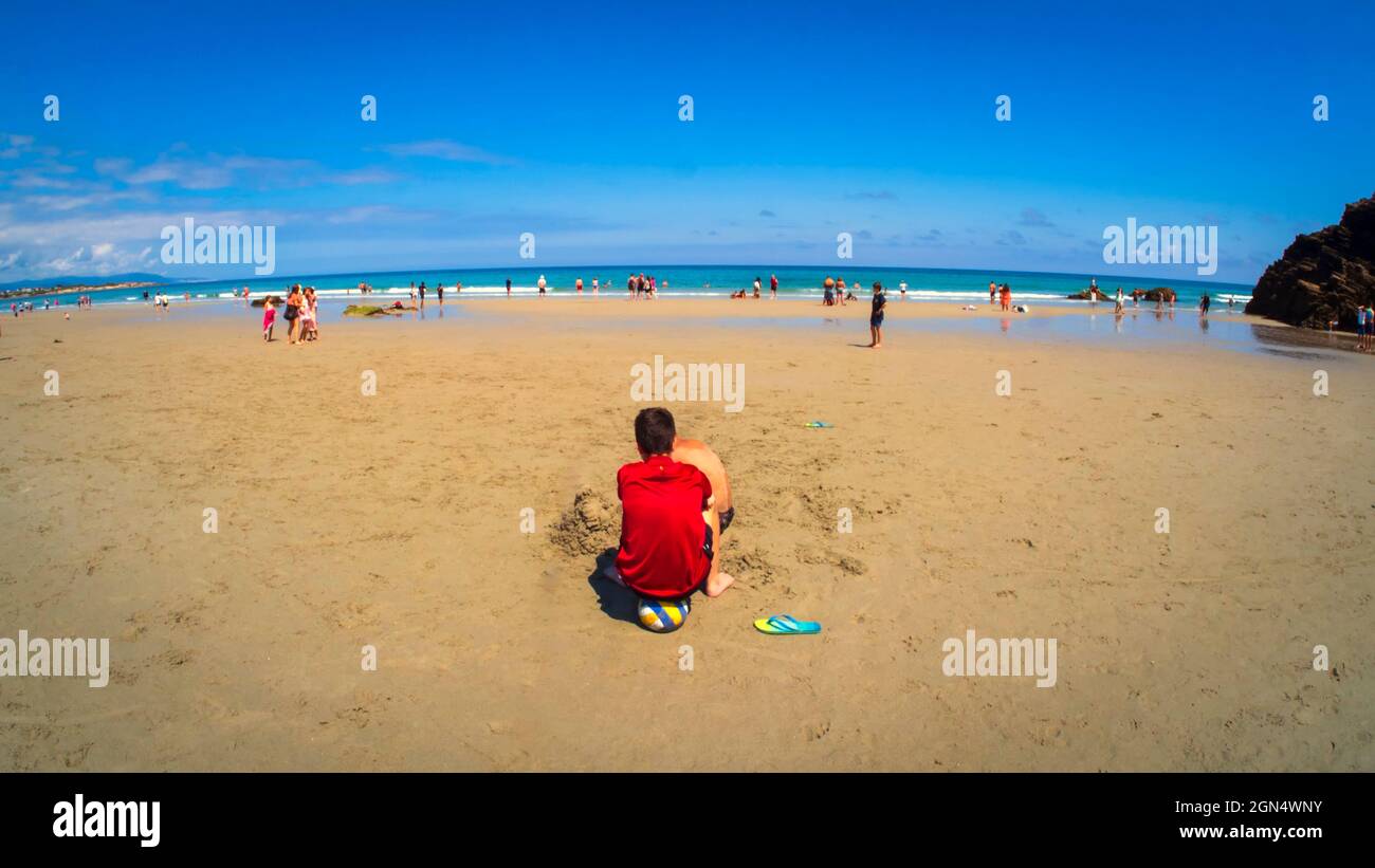 A boy in a red T-shirt plays with the sand on the beach of Las Catedrales on a blue-sky summer day. Stock Photo