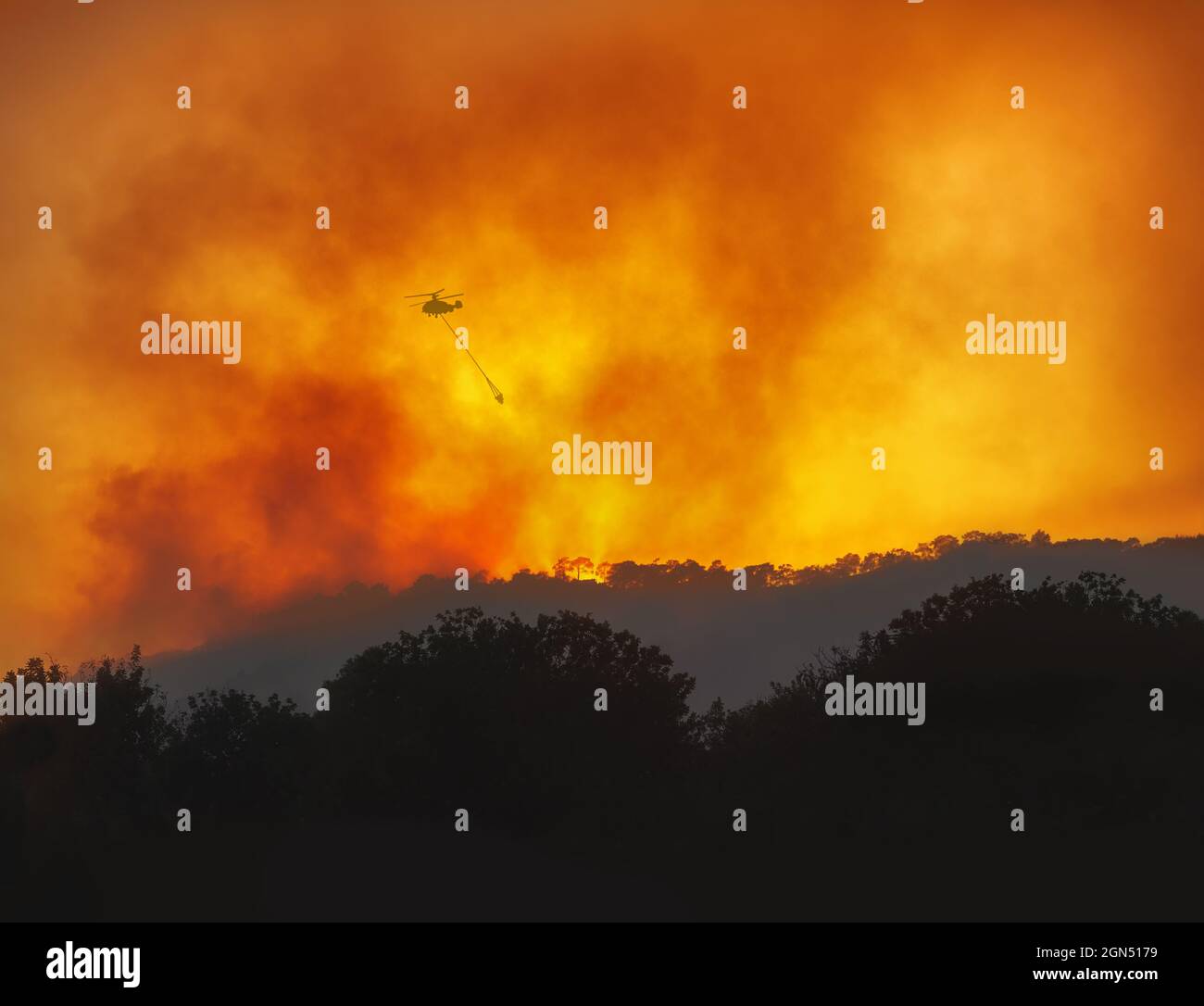 Helicopter with water bucket fighting forest wildfire at night, dramatic landscape with red sky and heavy smoke Stock Photo