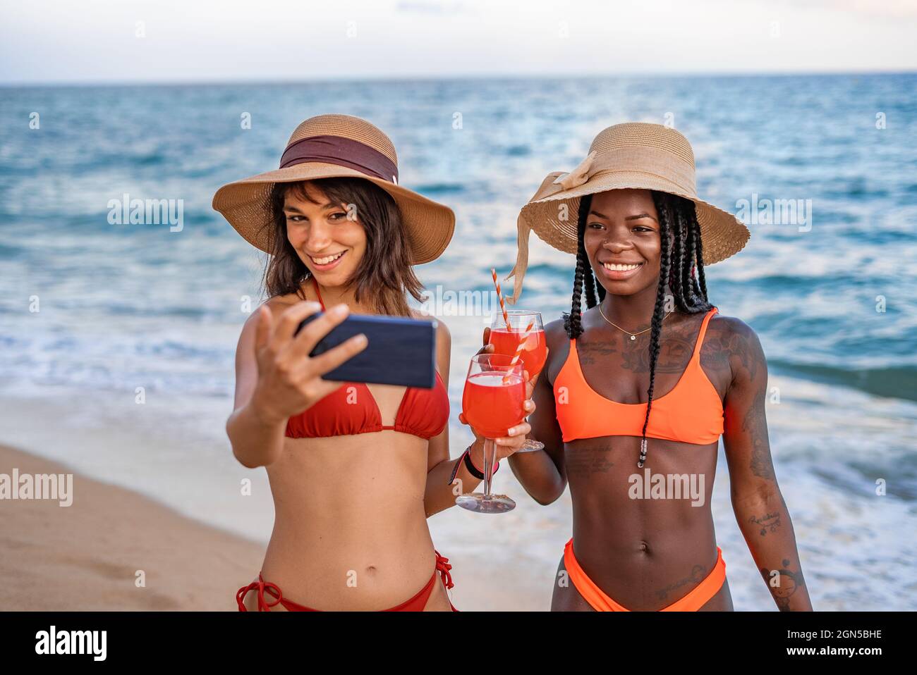 Young slim multiracial ladies in bikinis and hats clinking glasses with beverages and taking selfie on mobile phone while enjoying summer holidays on beach Stock Photo