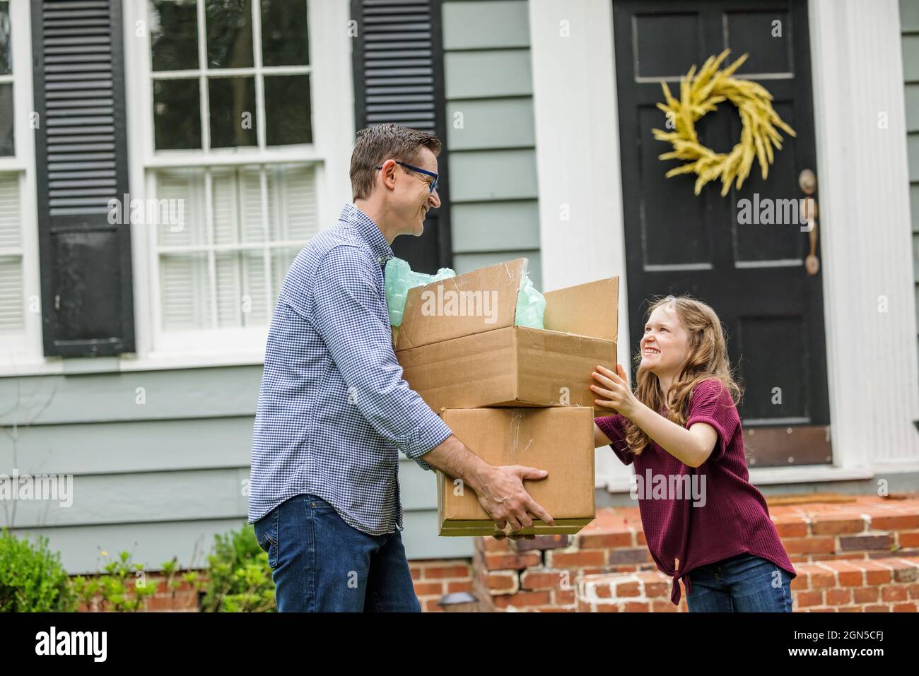 A father and daughter holding moving boxes outside a small blue cottage house getting ready for a move Stock Photo