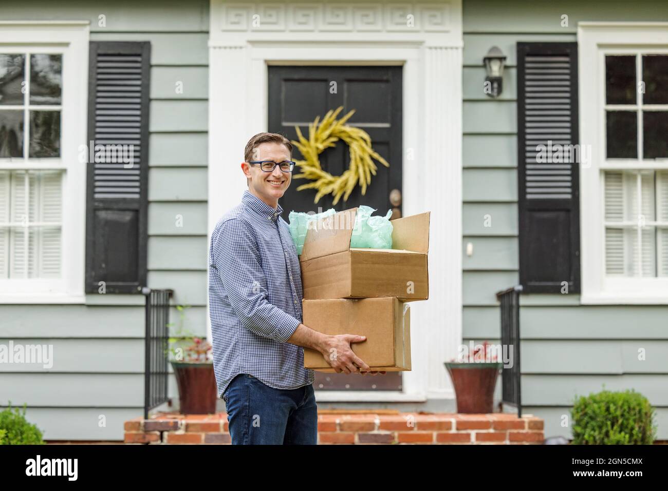 A man and father holding moving boxes outside a small blue cottage house getting ready to move into his new house Stock Photo