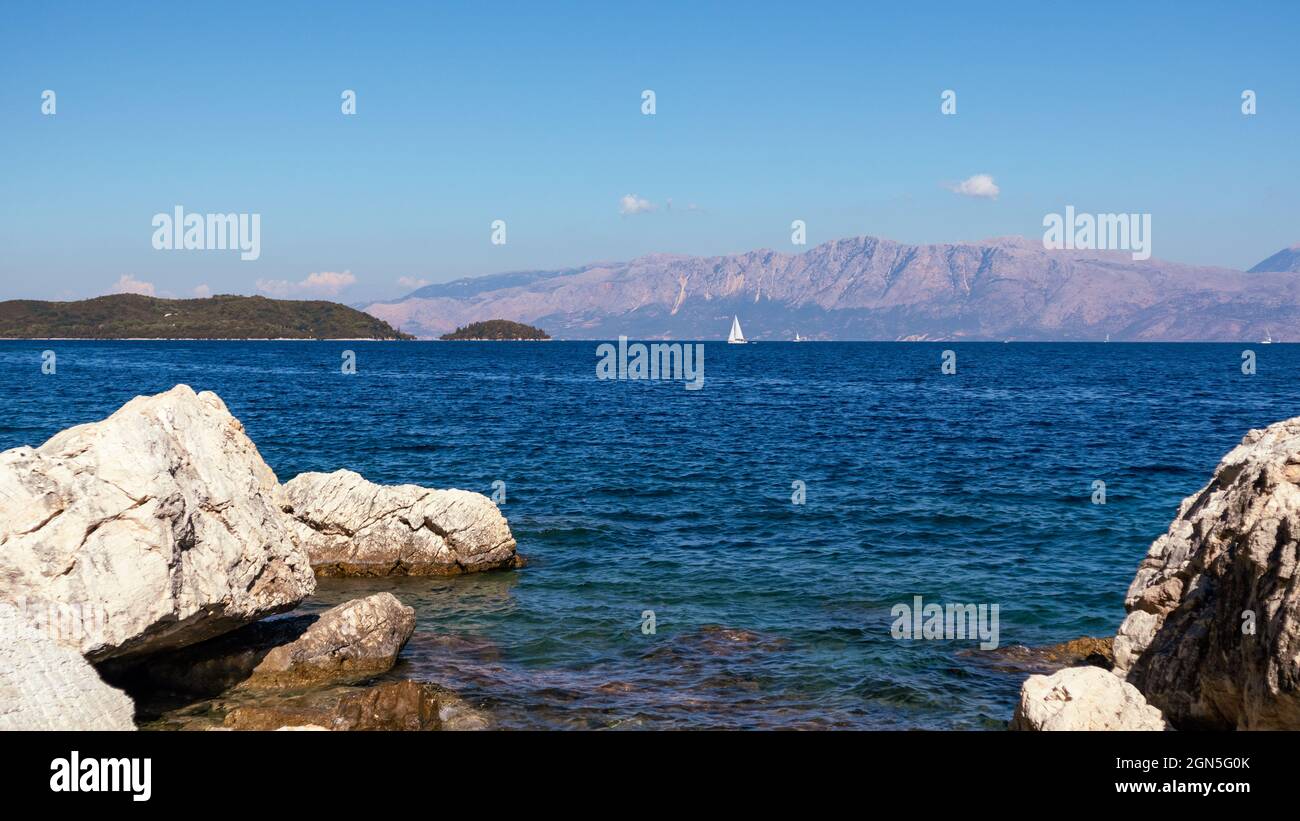 Big rocks in blue water on coast with white sail yacht and mountains on horizon, Lefkada island in Greece. Vivid wild nature travel to Ionian Sea Stock Photo