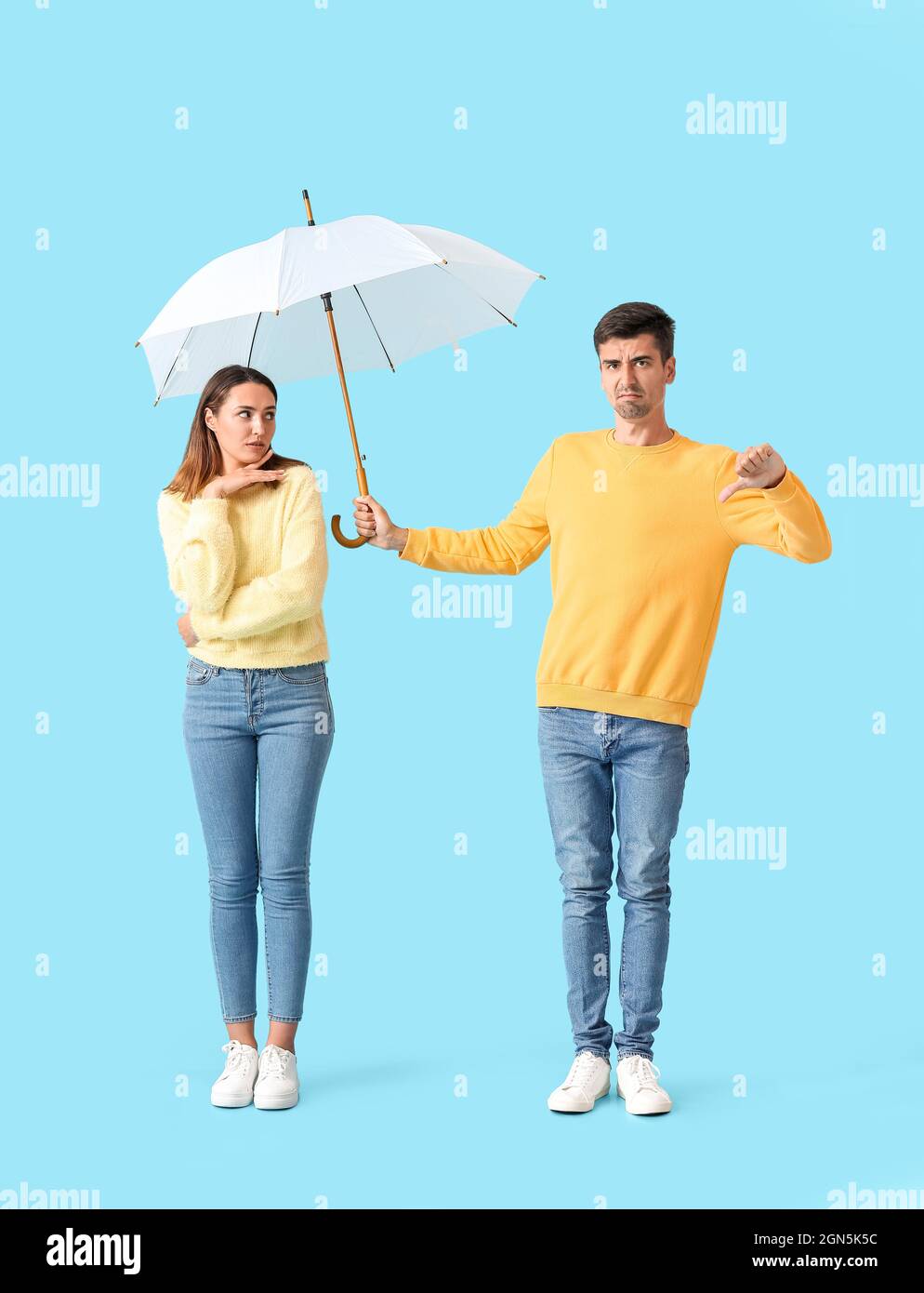 Displeased guy sharing umbrella with young woman on color background Stock Photo