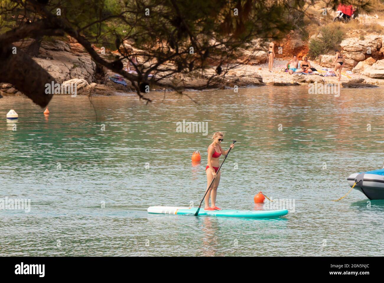 Kosirina, Murter, Croatia - August 24, 2021: Young woman in red swimsuit paddling on stand up board, on the beach, through pine tree Stock Photo