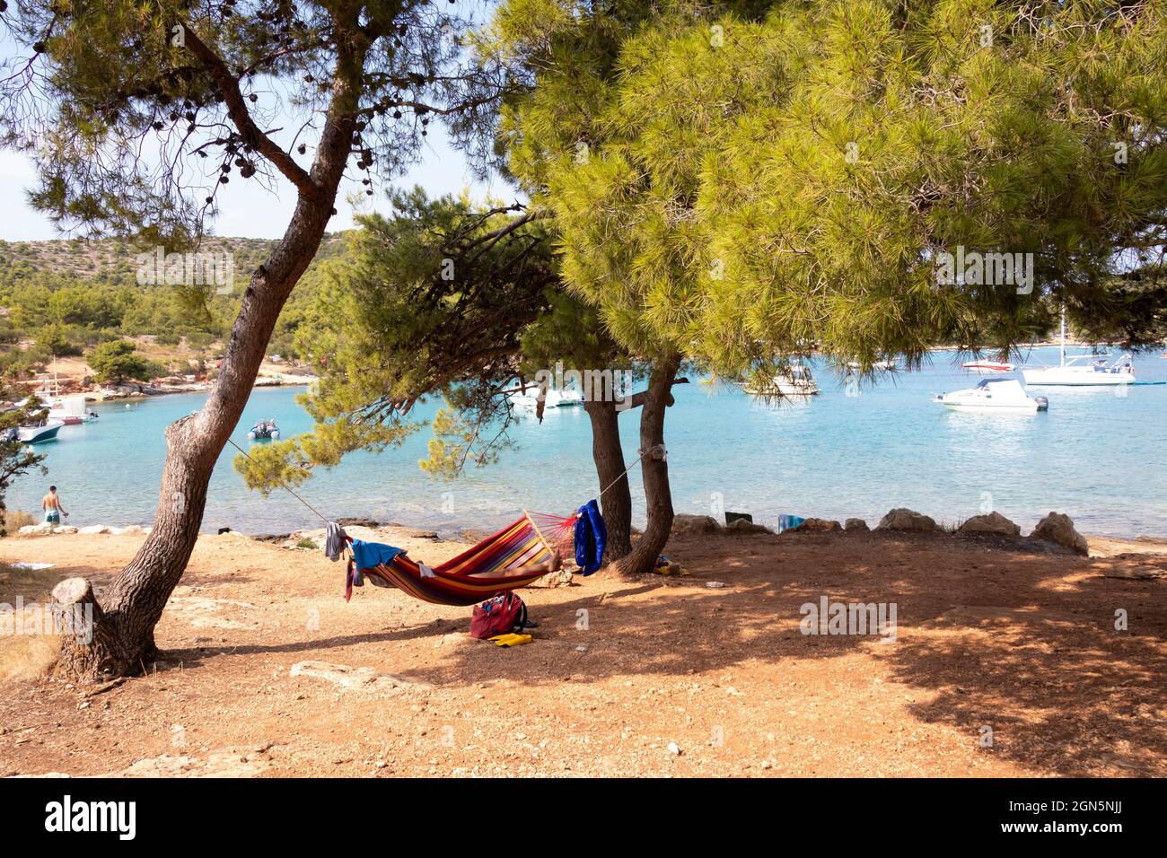 Kosirina, Murter, Croatia - August 24, 2021: Person lying in colorful hammock in the shade of pine trees on the beach and boats moored in a bay in per Stock Photo