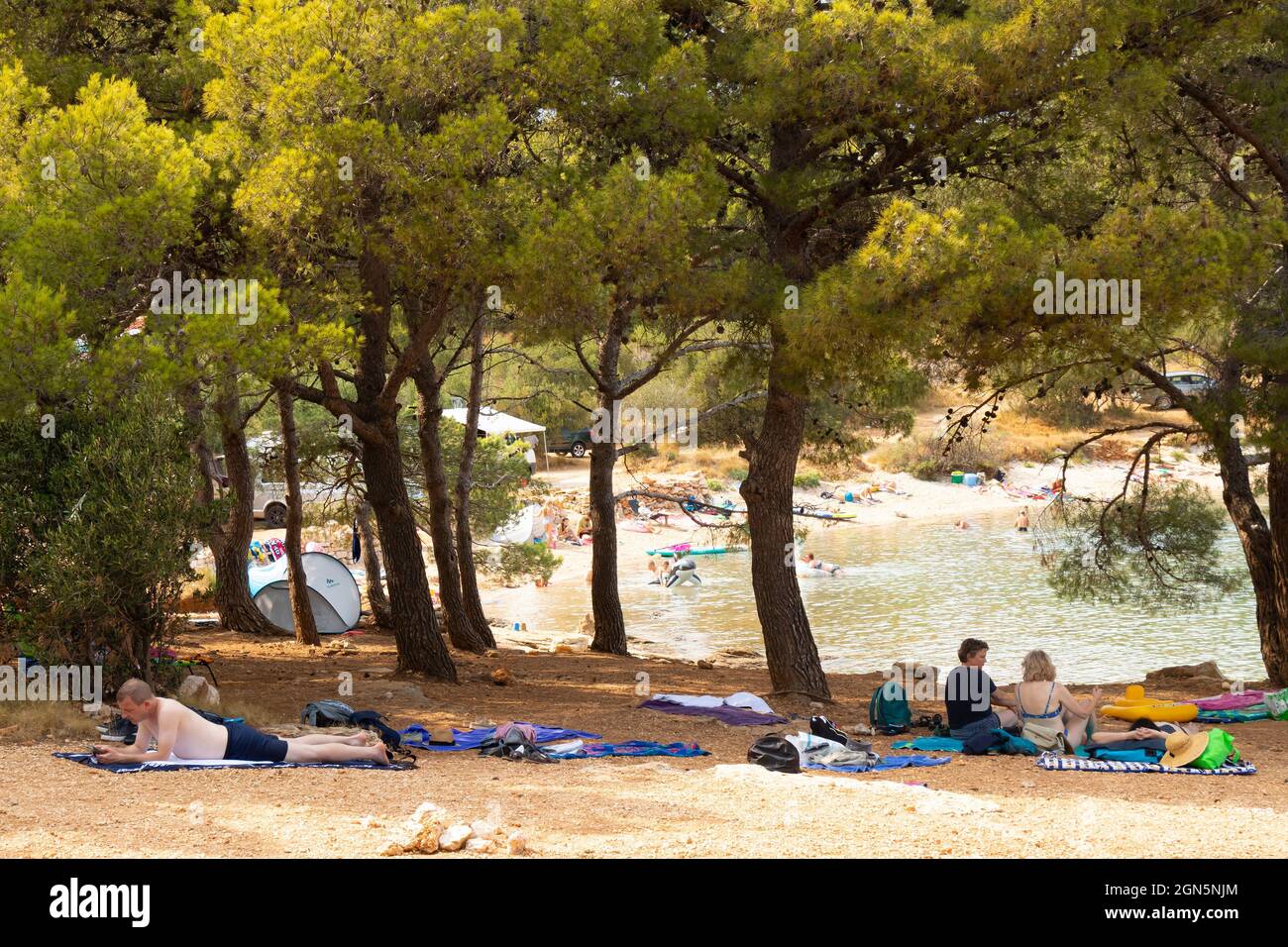 Kosirina, Murter, Croatia - August 24, 2021: People resting in the shade of pine trees on the beach and others swimming in a sea Stock Photo