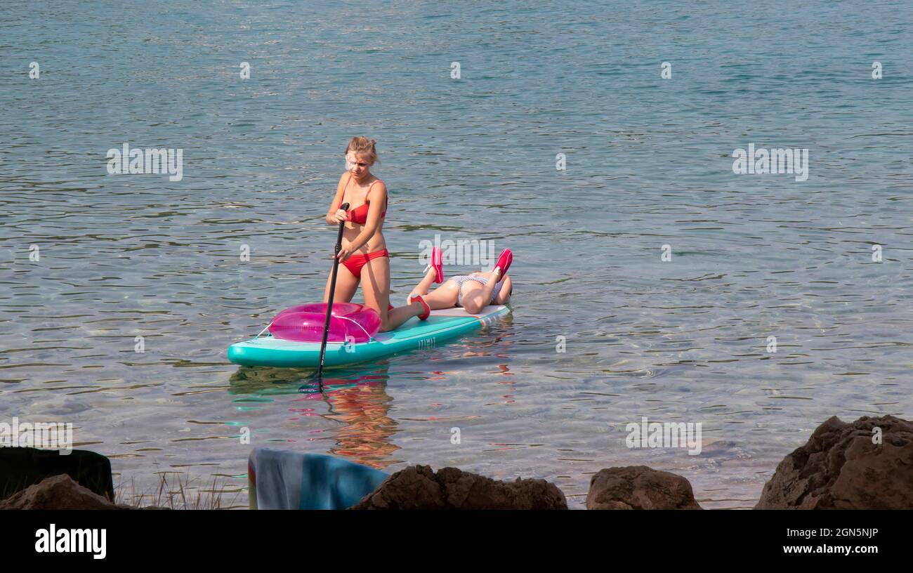 Kosirina, Murter, Croatia - August 24, 2021: Young woman in red swimsuit paddling on stand up board with little girl playing, on the rocky beach water Stock Photo