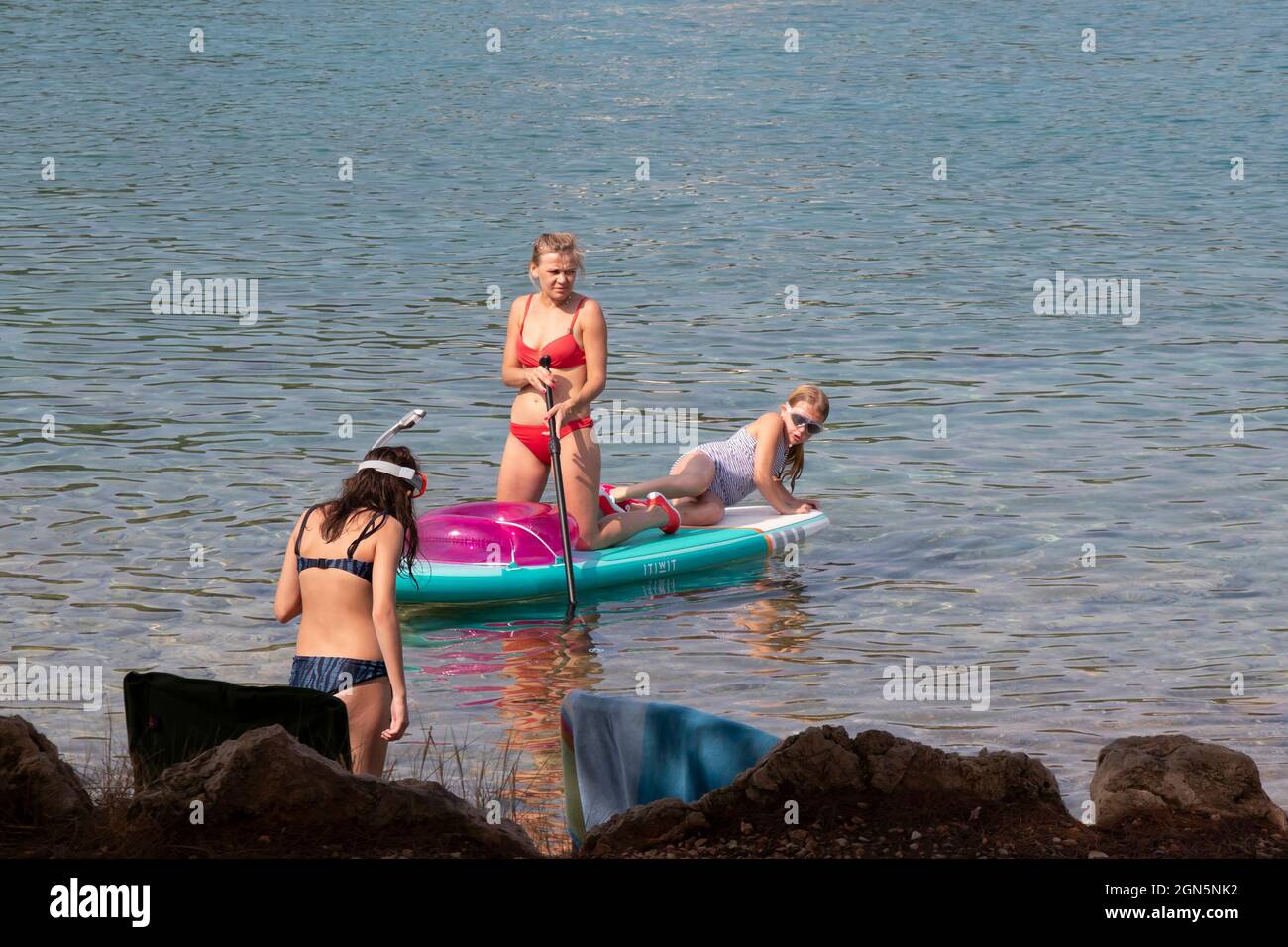 Kosirina, Murter, Croatia - August 24, 2021: Young woman in red swimsuit paddling on stand up board with girls playing, on the rocky beach waters Stock Photo