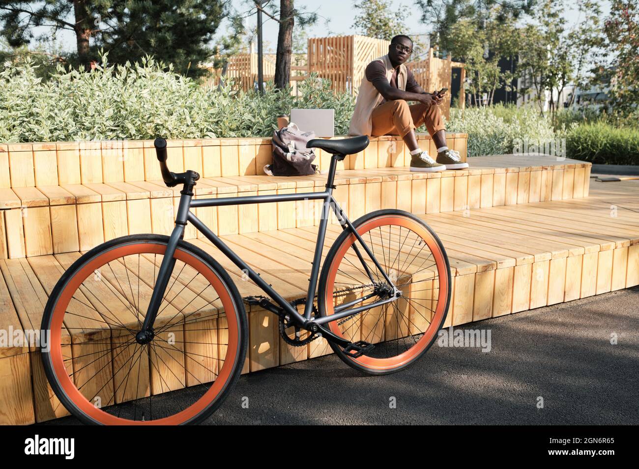 Horizontal selective focus shot of young African American man sitting on bench in modern city park looking at his bicycle parked nearby Stock Photo