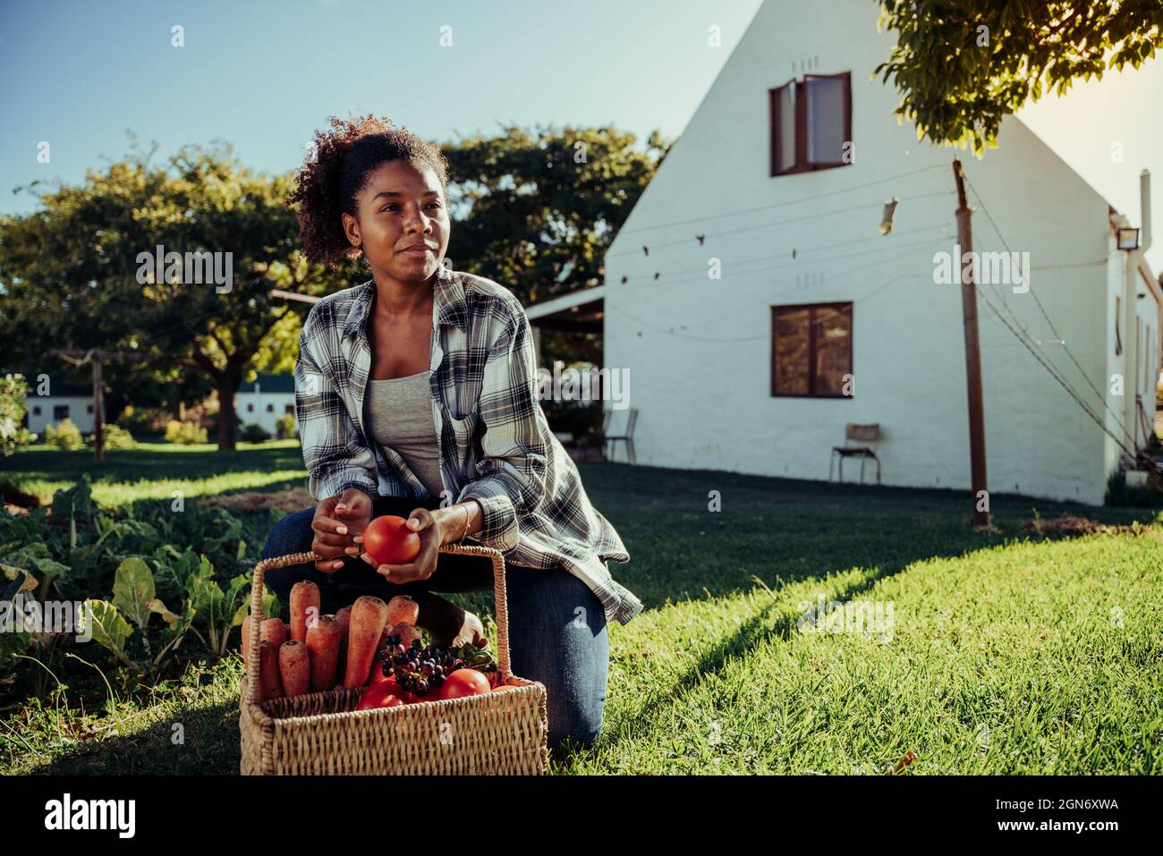 Mixed race female farmer crouching down in farm village grass smiling after successful harvest holding fresh vegetables  Stock Photo