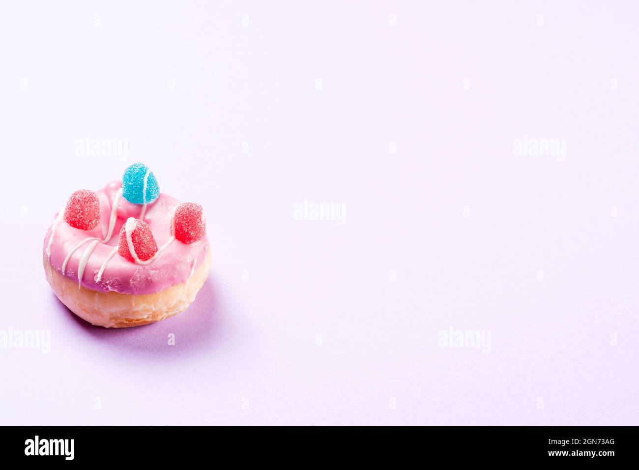 Photograph of 1 pink donuts decorated with jelly beans and drawn with white chocolate.The photo is taken in horizontal format on a lilac background an Stock Photo