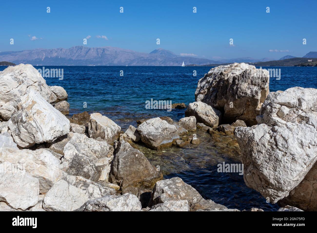 Big rocks in blue water on coast with sail yacht and mountains on horizon, Lefkada island in Greece. Summer vivid nature travel to Ionian Sea Stock Photo