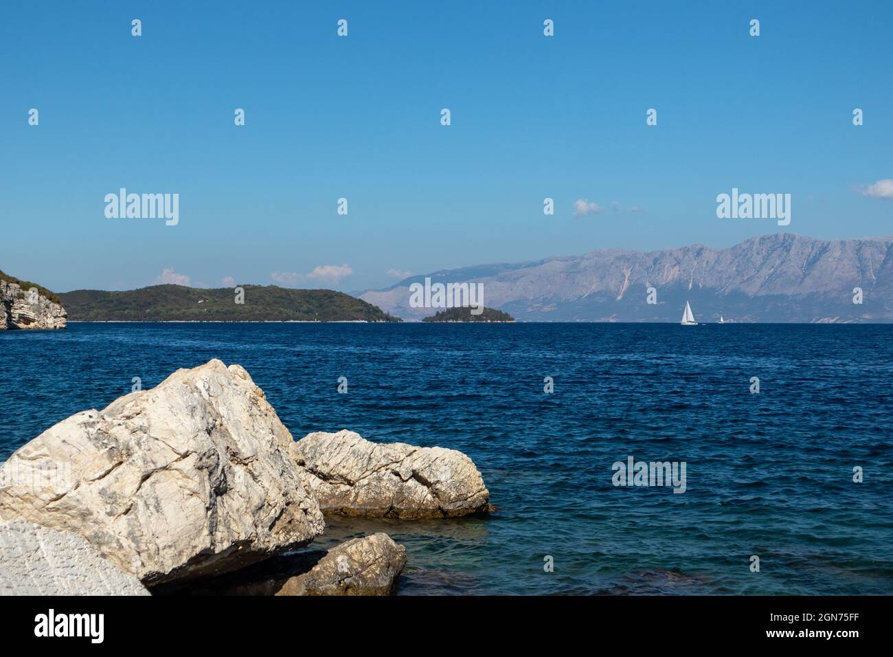 Big rocks in blue water on coast with white sail yacht and mountains on horizon, Lefkada island in Greece. Summer vivid nature travel to Ionian Sea Stock Photo