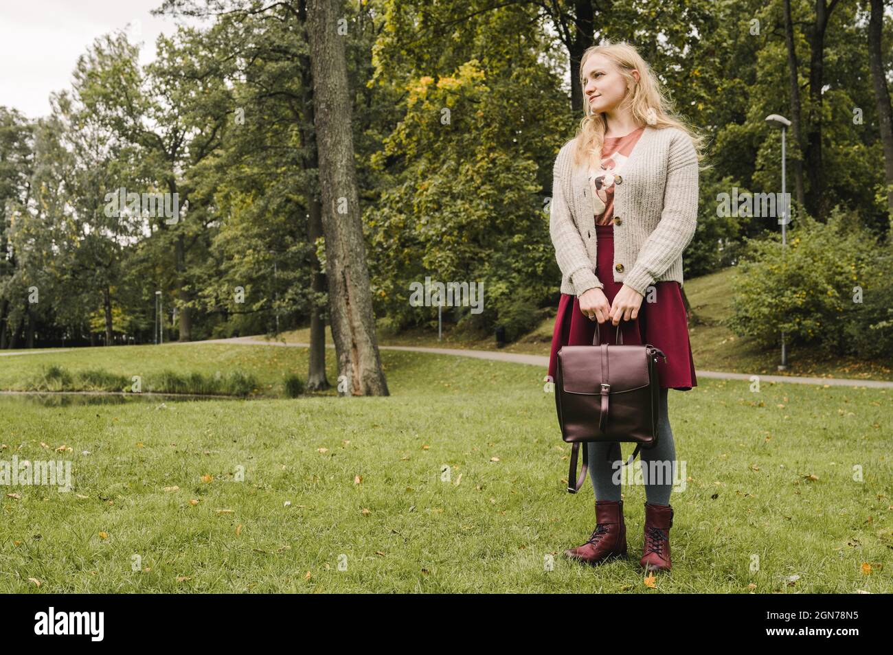 Young woman in knitted jacket, grey tights and skirt. Stylish burgundy shoes.  Leather bag in the arms. Green grass. Autumn fashion Stock Photo - Alamy