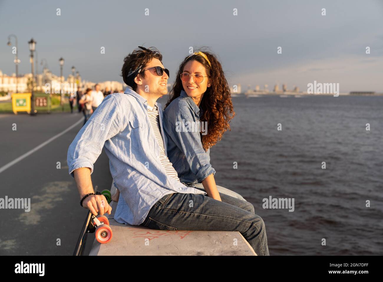 Stylish couple sit on pier at seaside enjoy sunset. Happy man and woman looking at river or sea Stock Photo