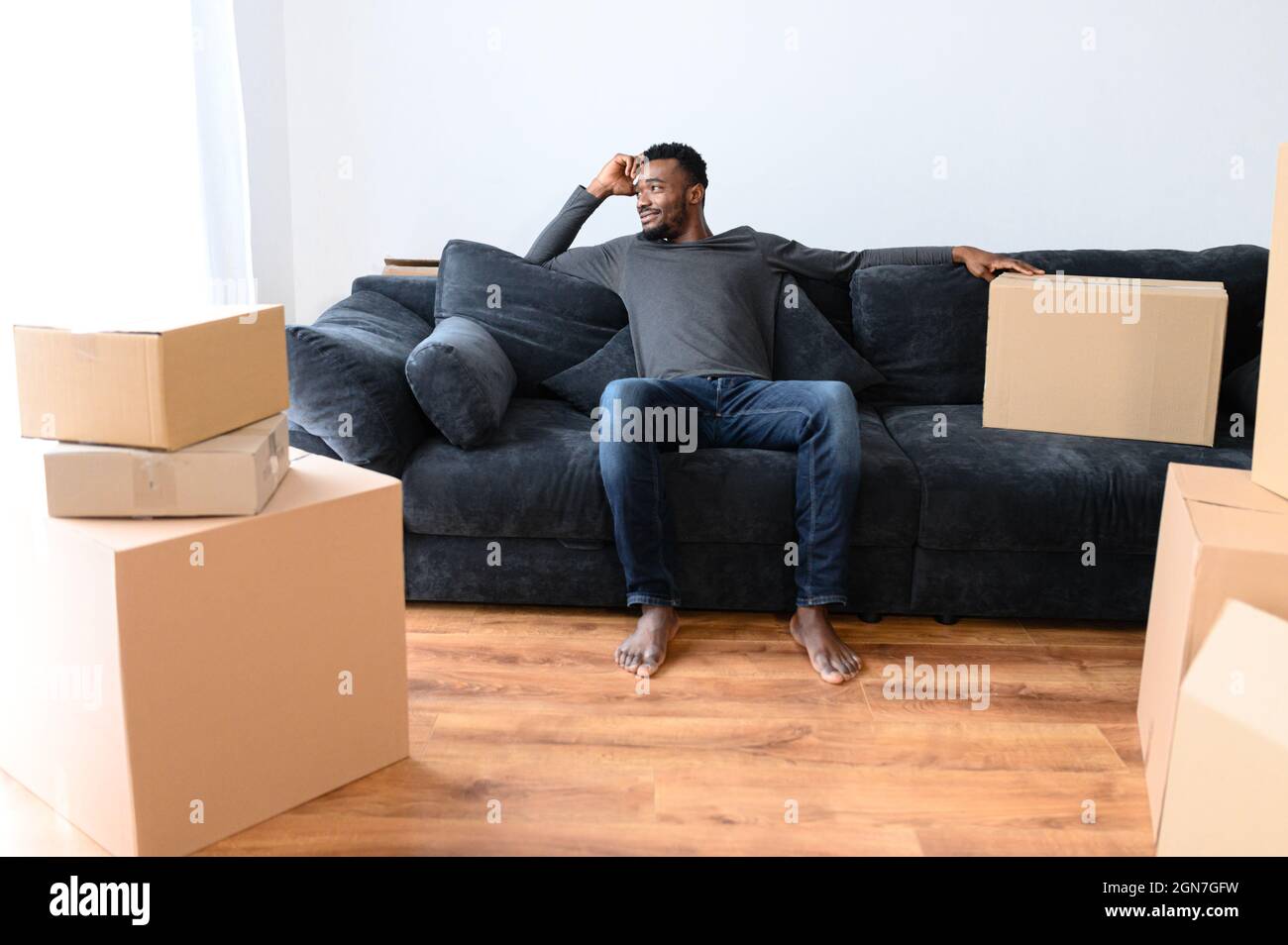Satisfied African American Guy Sitting On The Comfortable Couch And Looks Away Dreamly Among