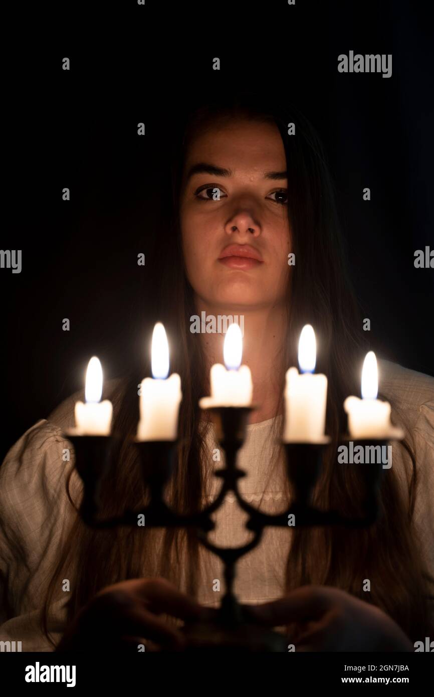 A young girl in an old white dress holding a candleabra in her hand and staring in to the camera. Dark background. Focus is on her face. Stock Photo