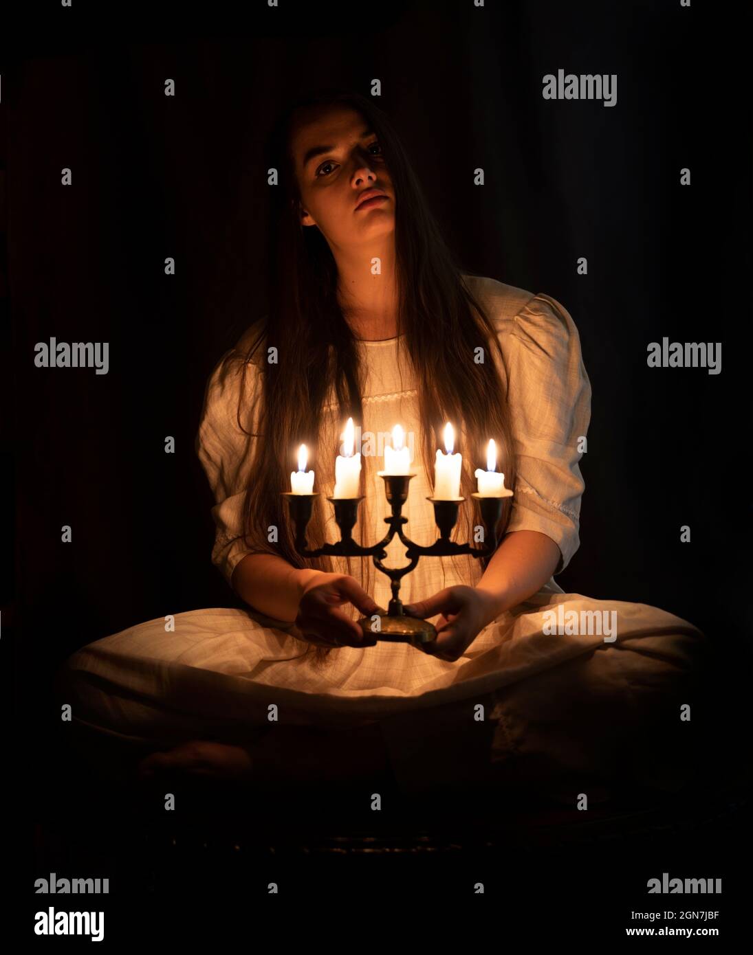 A young girl in an old white dress sitting and holding a candleabra in her hands. Dark background. Scary horror concept. Stock Photo