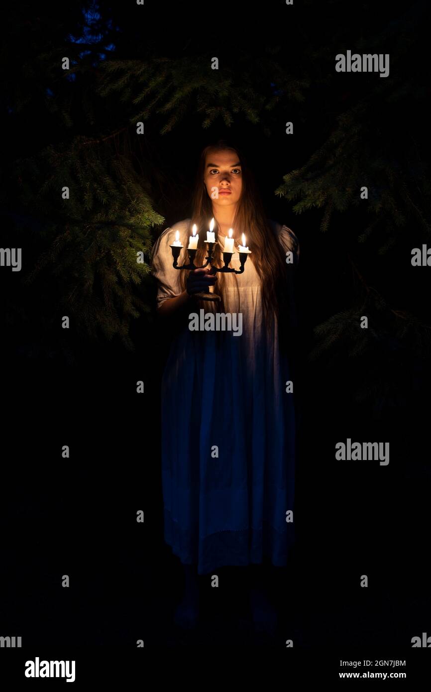 A young woman in the dark woods holding a candelabrum. Wearing an old long white dress. Stock Photo