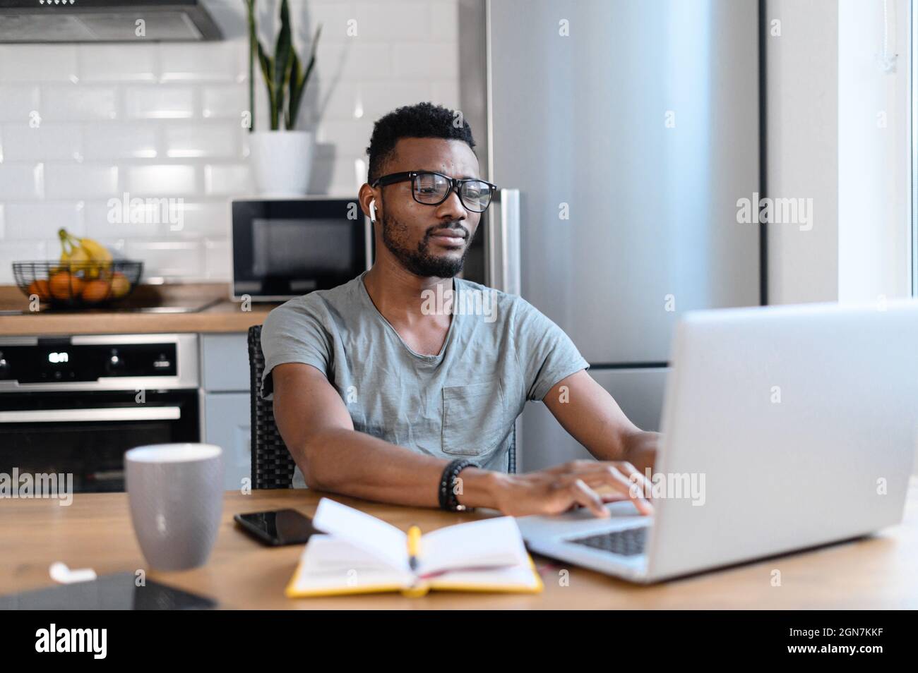 Concentrated African-American guy is working with a laptop sitting in the kitchen. Black man in casual wear and eyeglasses is typing on the keyboard, answering email, communicates with the customer Stock Photo