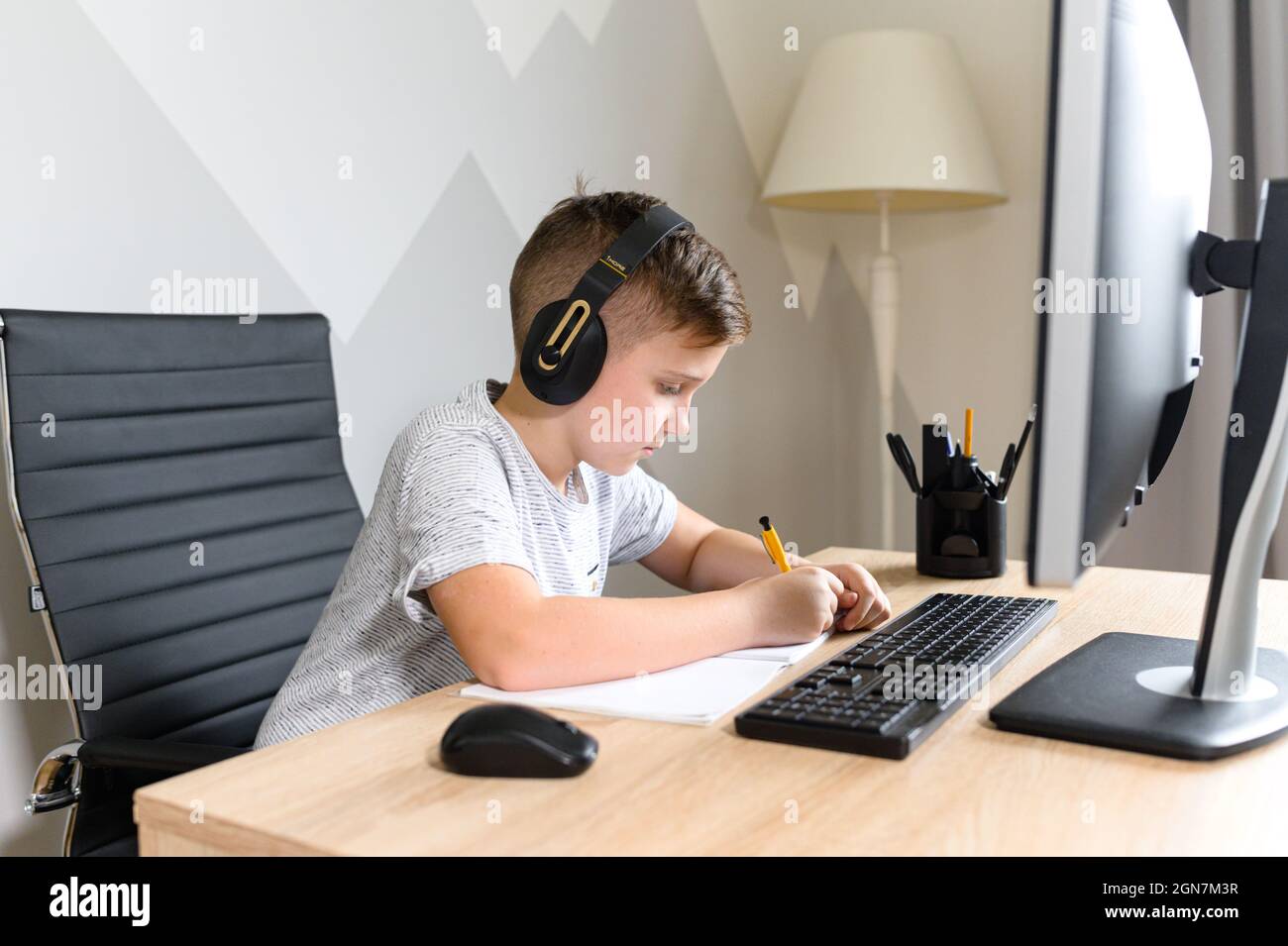 Schooler boy wearing headphones is studying in front of the computer, takes notes, listening music and writing down with a pen, preparing to exam. E-learning, study at home online, homeschooling Stock Photo