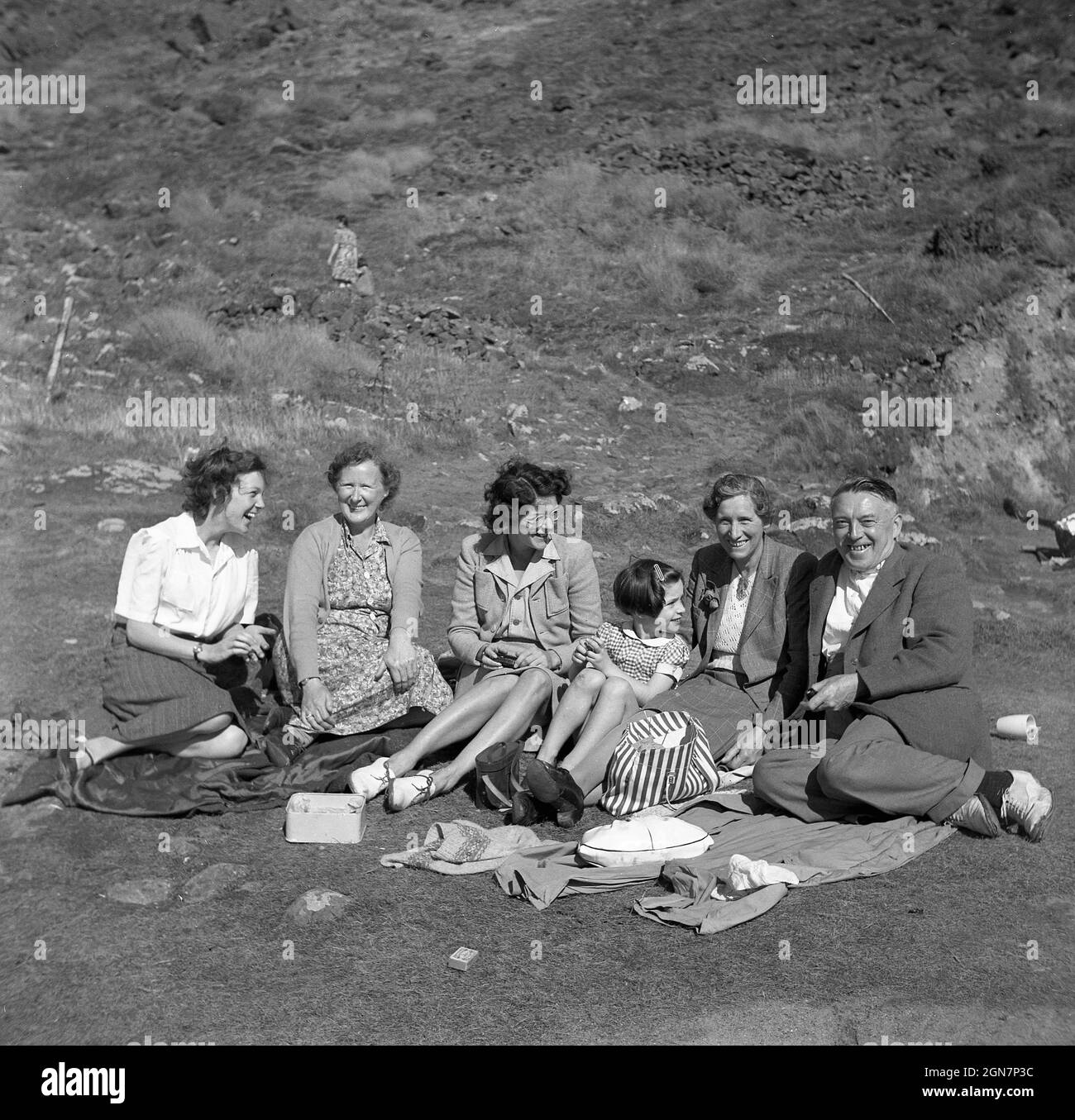 1950s, historical, four women, a young girl and elderrly man, probably the girl's grandfather sitting on their coats on a grassy hillside enjoying themselves together, having a laugh and having a light snack, maybe some sandwiches or cake, England, UK. Stock Photo