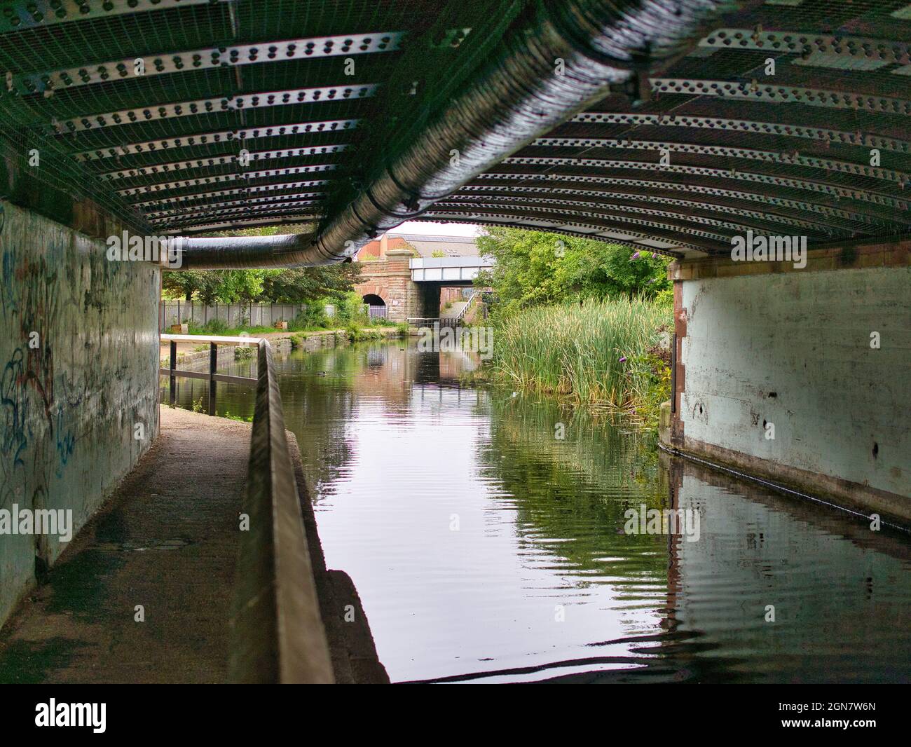 The darkened towpath of the Leeds Liverpool Canal as it passes under a rivetted metal bridge and pipework at the A5057. Stock Photo