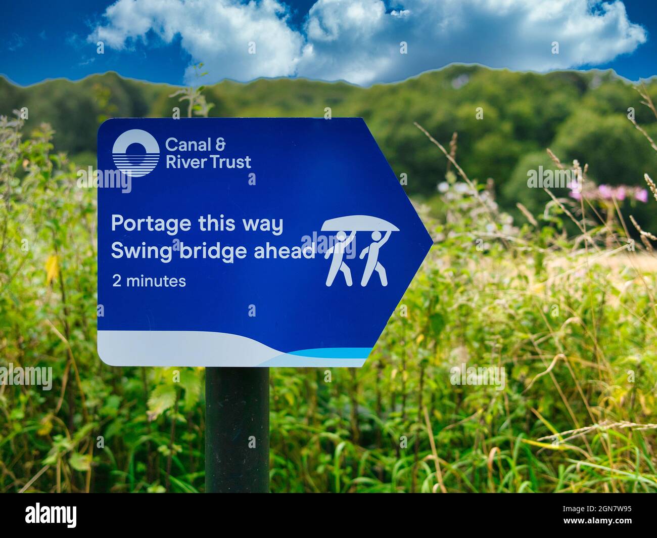 A blue Canal and River Trust sign on a canal towpath with white lettering advises those in rowing boats and canoes to carry their boat around a bridge Stock Photo