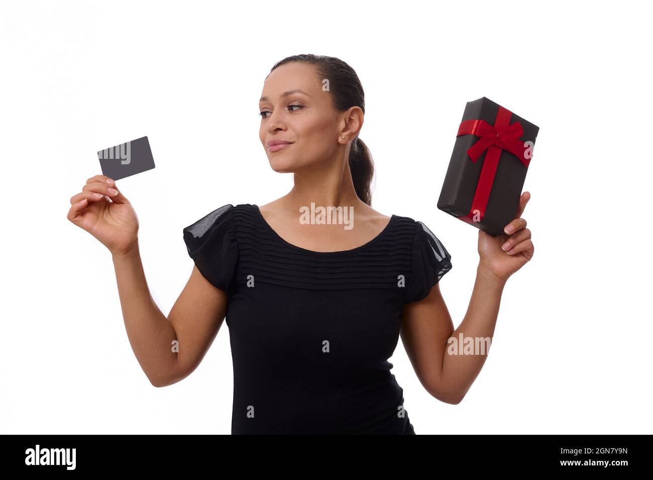 Beautiful mixed race African American woman looks at plastic credit or discount card in her hand and holds a black gift box with red ribbon in her oth Stock Photo
