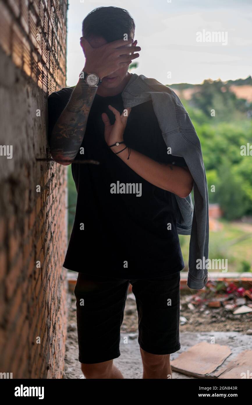 Portrait of young handsome Caucasian man in black casual clothes, covering his face with hand. Holding denim jacket, Full arm tattoo Stock Photo