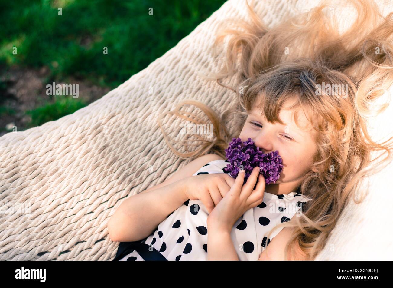 beautiful blond girl with long blond hair lying in white hammock and smelling lilac flower Stock Photo