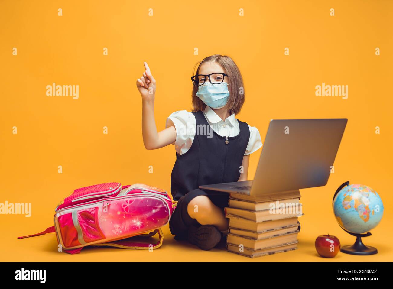 Schoolgirl in mask sits behind a stack of books and laptop raises index ...