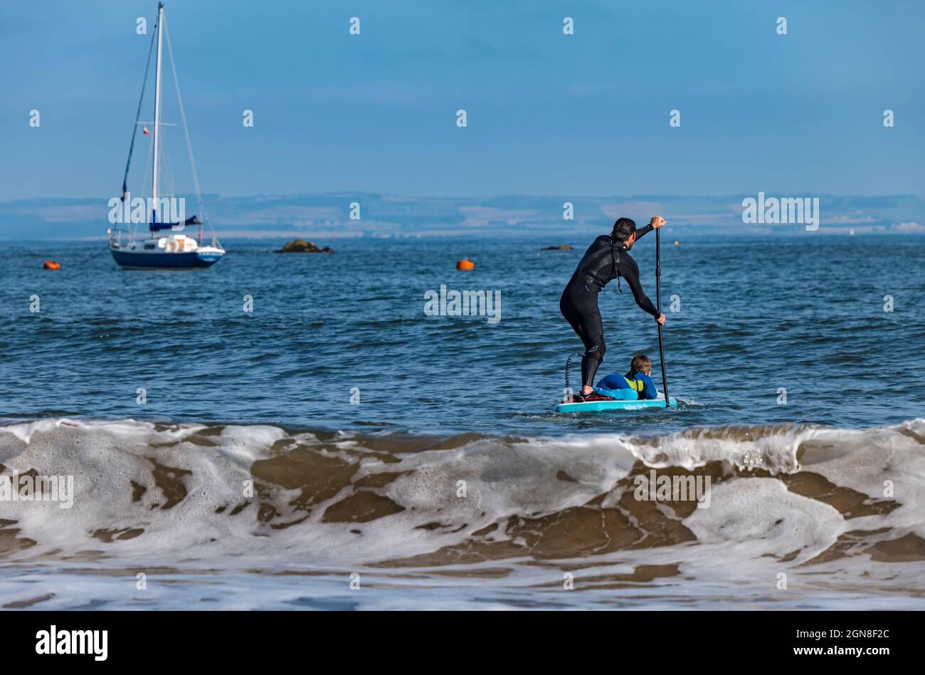 Man and boy paddle boarding in waves on beach in sunshine, North Berwick, East Lothian, Scotland, UK Stock Photo
