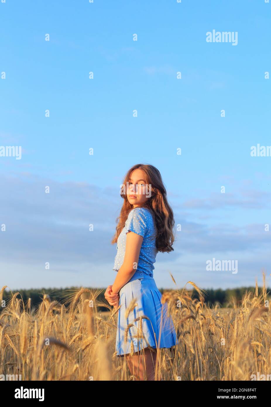 Happy young girl is smiling at wheat field, touching ears of wheat with her hand. Beautiful teenager enjoying nature in warm sunshine in a wheat field Stock Photo