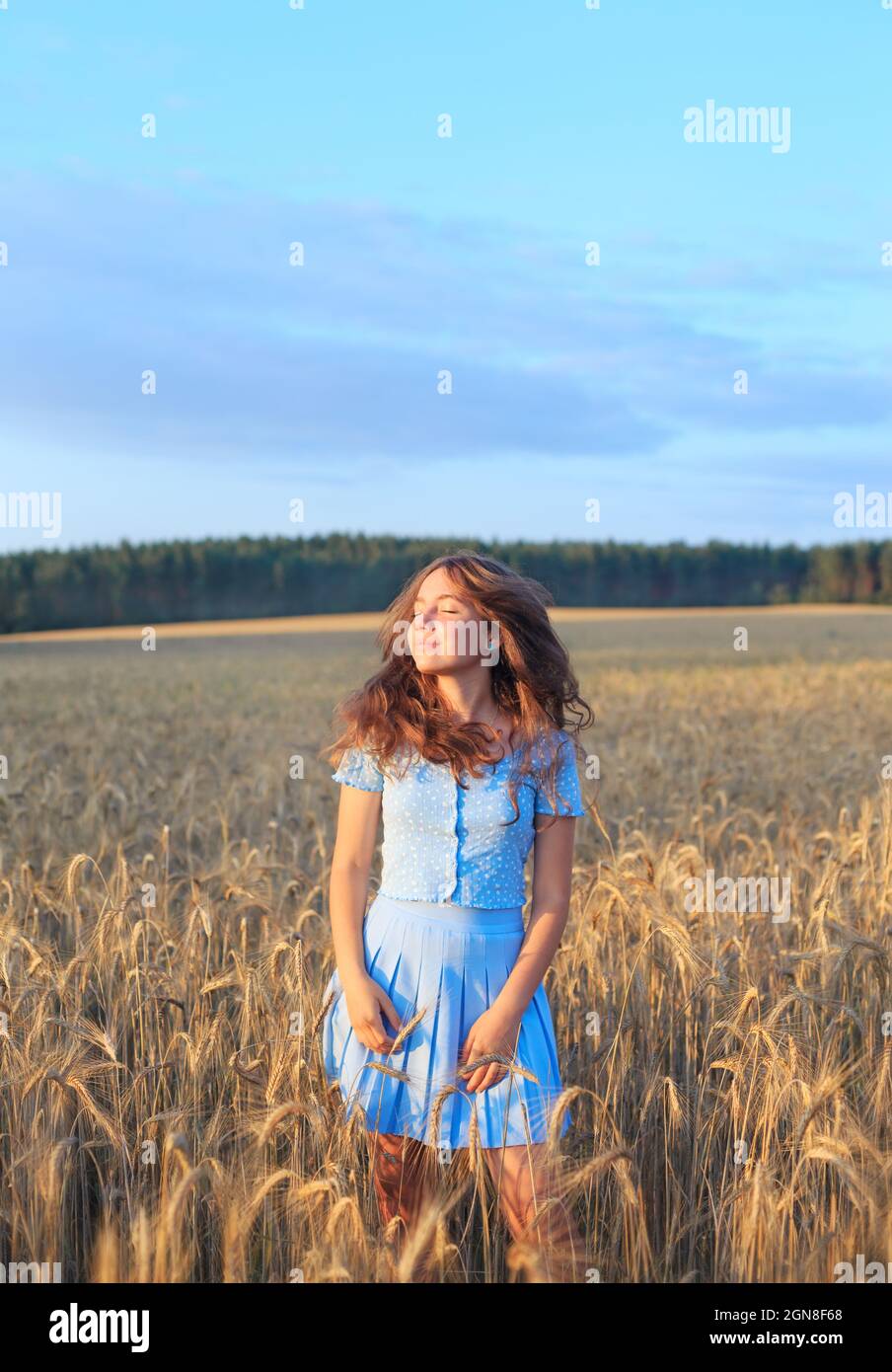 Happy teen girl is smiling at wheat field, touching ears of wheat with her hand. Beautiful teenager enjoying nature in warm sunshine in a wheat field Stock Photo